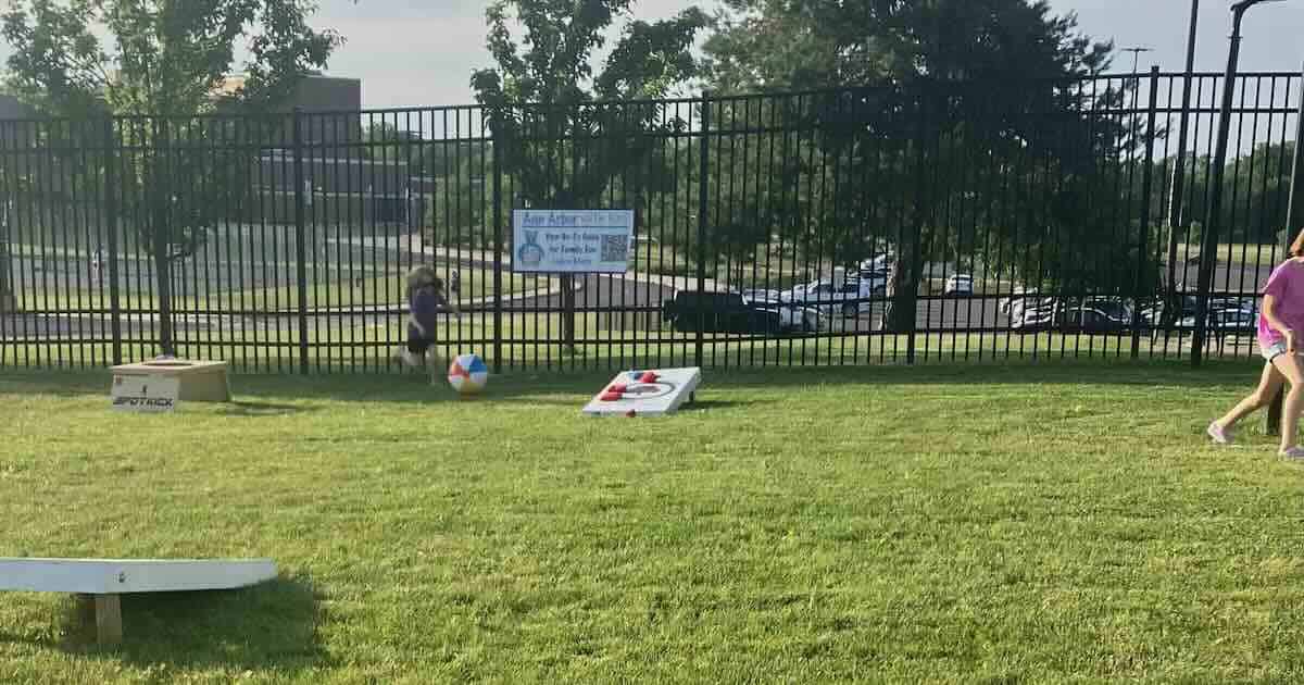AFC Ann Arbor Kids Area - Corn Hole, Spot Kick frame, child kicking a beach ball in front of a black metal fence with an Ann Arbor with Kids banner on it. Through the fence you can see Pioneer High School and the parking lot.