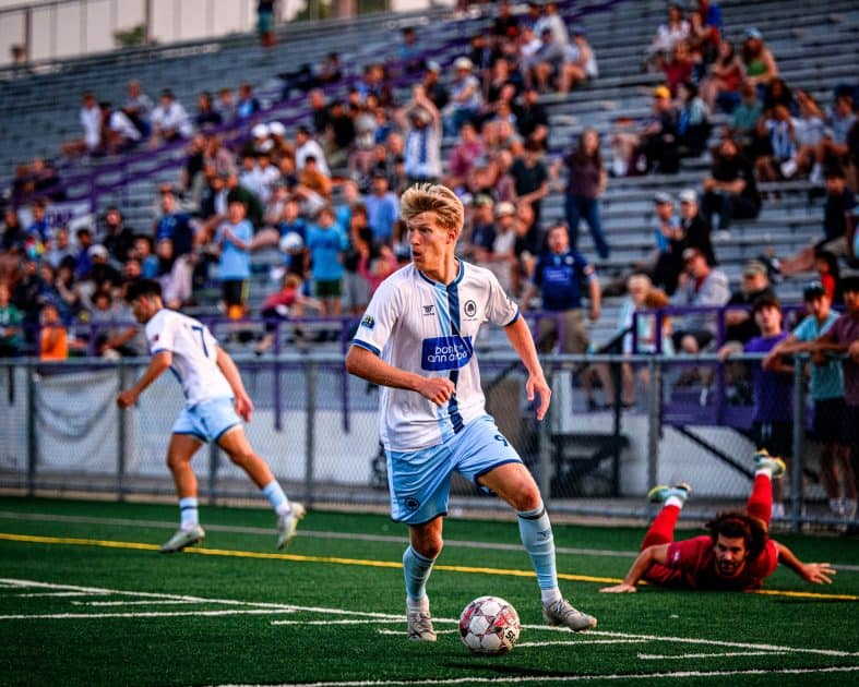 AFC Ann Arbor game. AFC Player with ball. Opposing team player laying on ground while another AFC AFC player heads in opposite direction. Plus fans cheering in stands