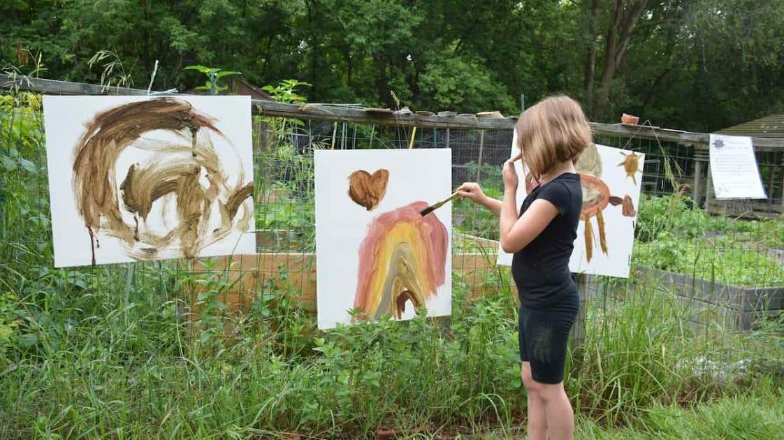 Child painting rainbow on a canvas hung on a garden fence. Other paintings hang nearby
