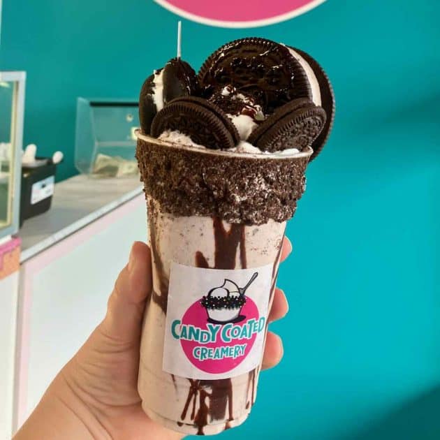 Milkshake held up in front of teal wall. Milkshake in a plastic cup with a fudge rim with cookie crumbs and topped with a large Oreo ice cream sandwich, and other cookies