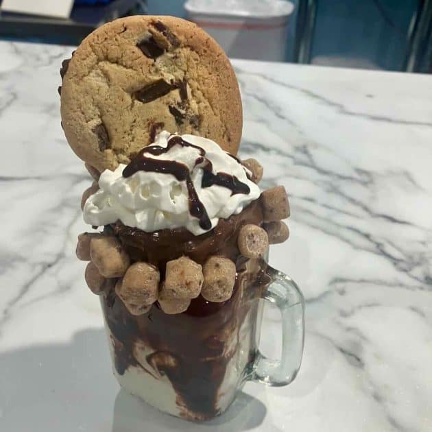 Chocolate milkshake with edge of rim coated in fudge with cookie dough bites encircling the rim, shake is topped with whipped cream, chocolate syrup, and a large cookie