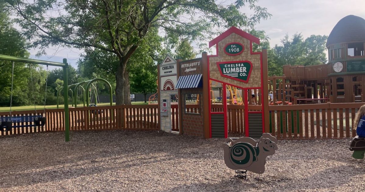 Timbertown tot area - includes facades of buildings - Chelsea Lumber, Heydlauff's Appliances, & Chelsea State Bank with the clock tower visible in the background -bouncers, swings, and tot lot fence.