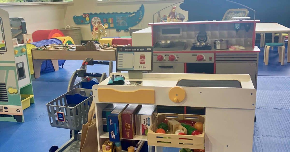 We Rock the Spectrum - imaginative play area highlighted by a market including a checkout counter. mini shopping carts are to the left, a kitchen is behind that, along with other preschool play items.