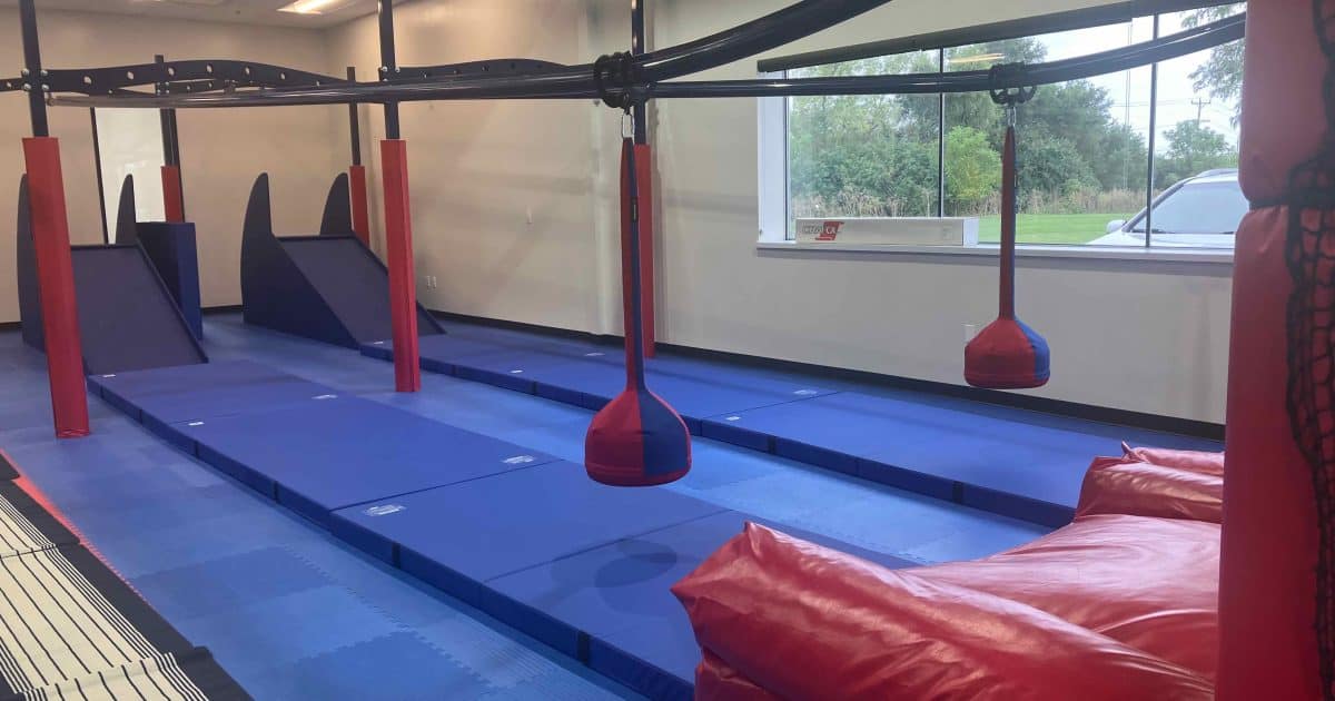 We Rock the Spectrum - Zipline - blue mats below an overhead bar holding a red & blue pendulum for kids to ride that goes from stairs at one end to a giant red inflatable pillow at the other end