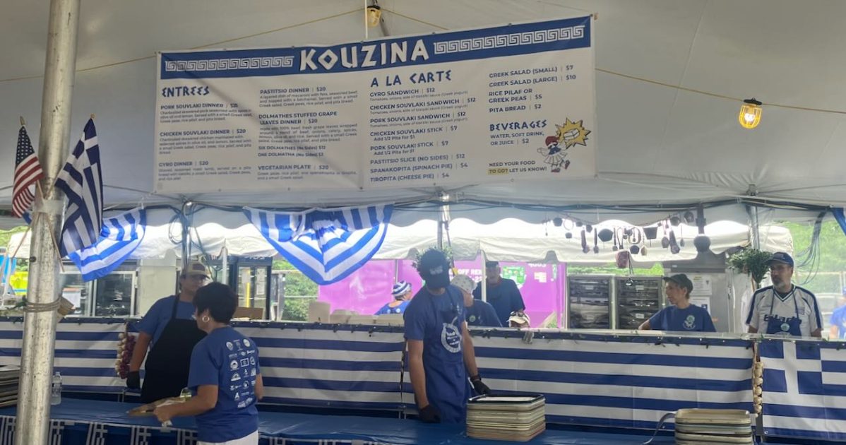 Ya'ssoo Greek Festival - Kouzina Food Stand - people serving and working in kitchen. Menu hangs above with Entrees & A La Carte Items